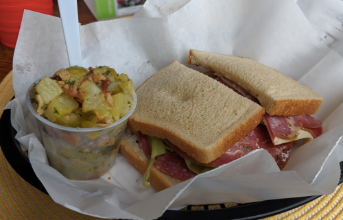 A sandwich with layers of meat and lettuce, served with a side of potato salad in a cup.