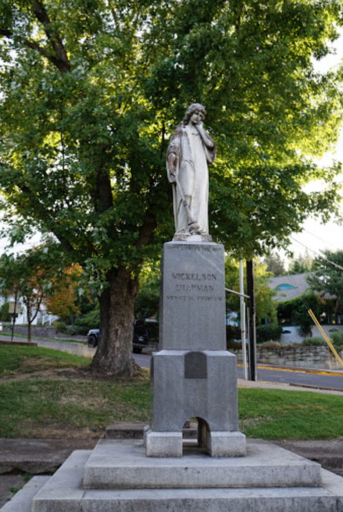 Statue of a figure holding a book, mounted on a stone pedestal, surrounded by trees and a street in the background.