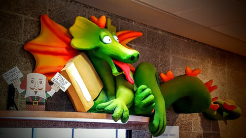 A whimsical green dragon with orange wings rests on a shelf, holding a book, surrounded by playful decorations.