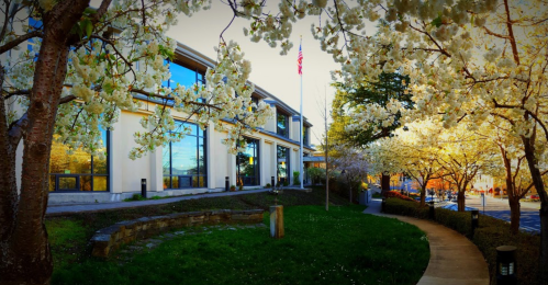 A modern building surrounded by blooming trees and greenery, with an American flag visible in the background.
