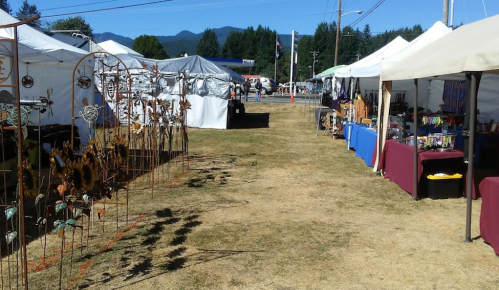 A sunny outdoor market with vendor tents on either side, showcasing crafts and decorations along a grassy path.