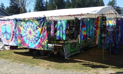A vibrant outdoor market stall displaying colorful tie-dye fabrics and clothing under a white tent.