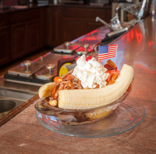 A banana split topped with whipped cream, nuts, and a cherry, served in a glass dish with a small American flag.