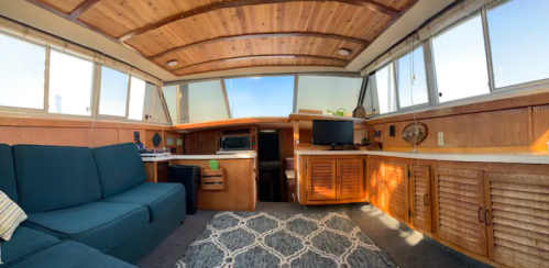 Cozy interior of a boat with wooden paneling, a sofa, and a small desk, featuring large windows for natural light.