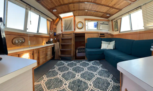 Cozy interior of a boat with wooden walls, a blue couch, a rug, and a small staircase leading to a sleeping area.