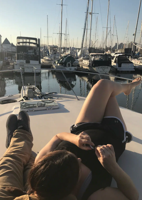 A person relaxes on a boat, with their feet up, surrounded by a marina filled with sailboats and a clear sky.