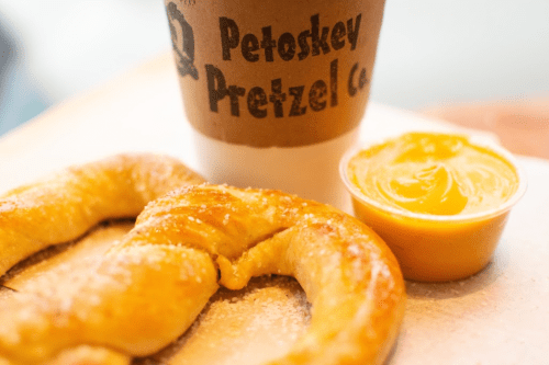A soft pretzel dusted with salt next to a cup of cheese sauce and a coffee cup from Petoskey Pretzel Co.