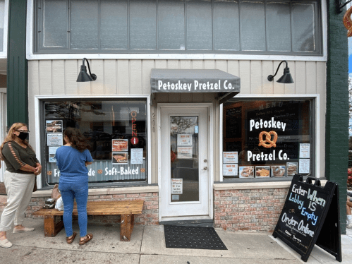 Exterior of Petoskey Pretzel Co. with a sign, menu, and two customers outside, one wearing a mask.