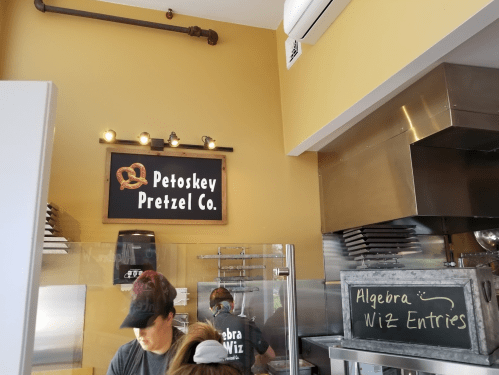 Interior of Petoskey Pretzel Co. with staff working and a chalkboard menu displaying "Algebra Wiz Entries."