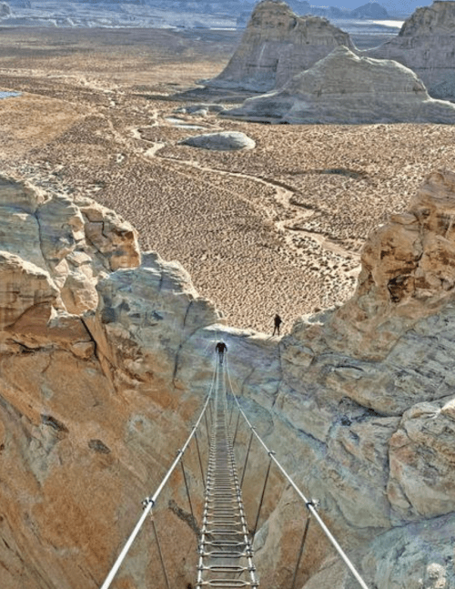 A narrow suspension bridge stretches across a deep canyon, with rocky cliffs and desert landscape in the background.