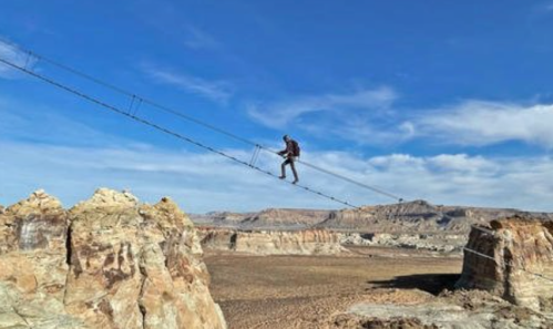 A person walks on a high rope bridge between rocky cliffs under a blue sky with scattered clouds.