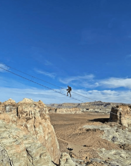 A person balances on a high wire between rocky cliffs, with a vast desert landscape and blue sky in the background.