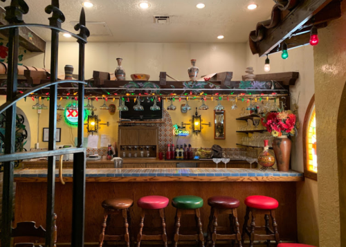A cozy bar with colorful lights, wooden stools, and a decorated counter featuring bottles and vibrant decor.