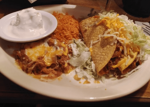 A plate of Mexican food featuring rice, refried beans with cheese, two tacos, lettuce, and a side of sour cream.