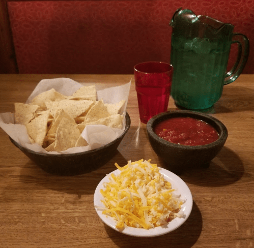 A bowl of tortilla chips with salsa, a small bowl of shredded cheese, and drinks on a wooden table.