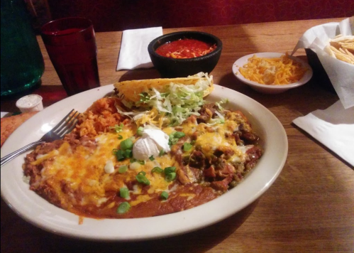 A plate of enchiladas topped with cheese and green onions, served with rice, a taco, salsa, and shredded cheese on the side.