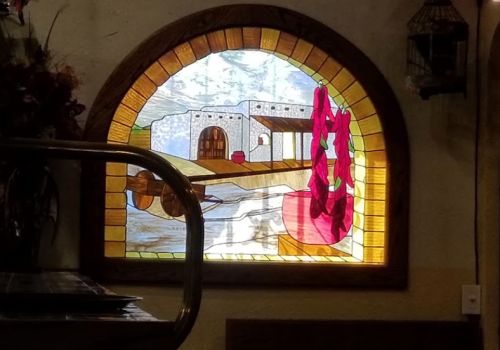 Stained glass window depicting a scenic view of a building by a body of water, framed in an arched wooden border.