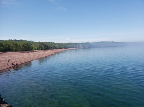 A serene beach scene with a sandy shore, calm blue water, and misty trees in the background under a clear sky.