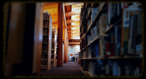 A view of a library aisle lined with bookshelves, featuring wooden beams and warm lighting.