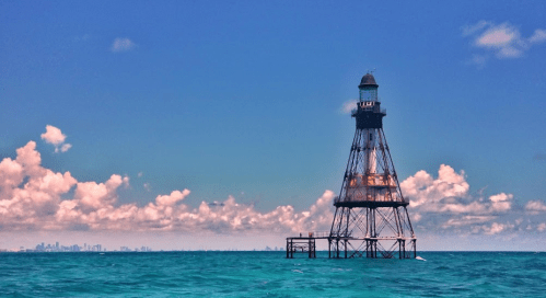 A tall lighthouse stands in the ocean, surrounded by water and clouds, with a city skyline visible in the distance.