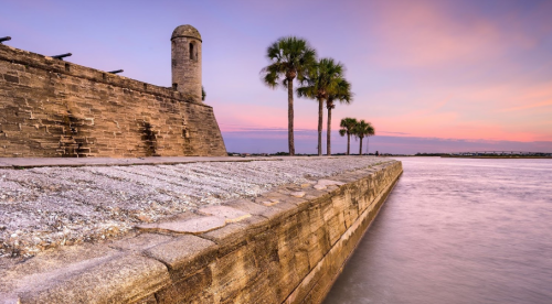 A stone wall with a turret beside a calm waterway, framed by palm trees under a colorful sunset sky.
