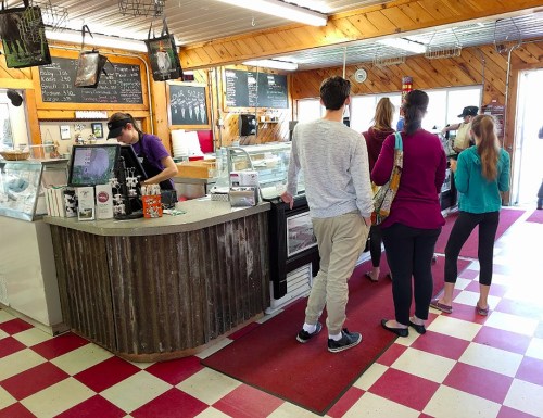 A busy ice cream shop with customers waiting in line at the counter, featuring a rustic interior and checkered floor.