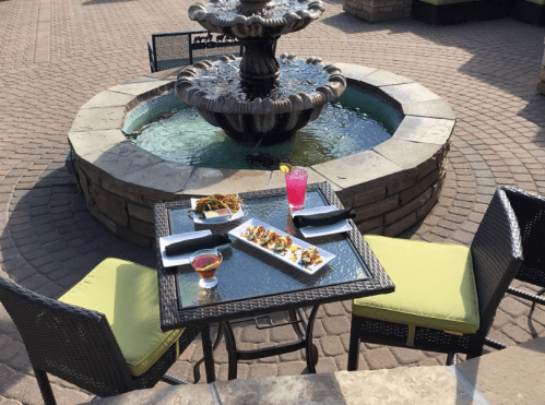 A table set with colorful dishes and drinks near a decorative fountain in a cozy outdoor dining area.