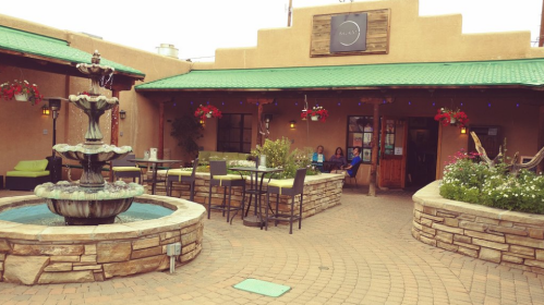 A charming courtyard with a fountain, seating, and hanging flowers outside a rustic building with a green roof.