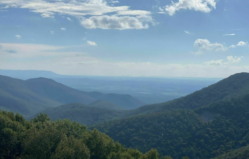 A scenic view of rolling mountains under a blue sky with scattered clouds, showcasing lush greenery and distant valleys.