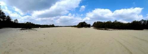 A wide sandy landscape under a blue sky with scattered clouds and patches of greenery in the distance.