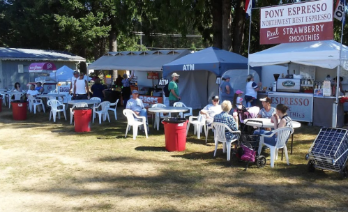 Outdoor scene with people sitting at tables near food stalls and an ATM, surrounded by trees and tents.