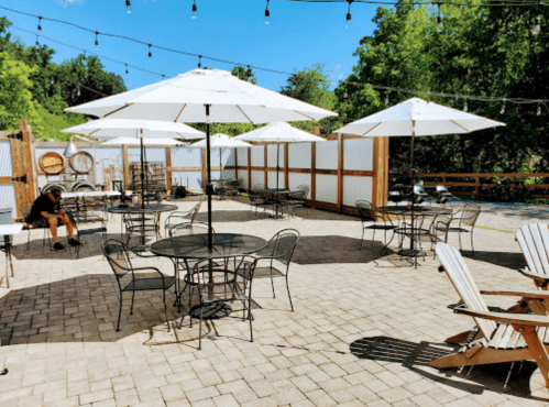Outdoor patio with tables, chairs, and umbrellas, surrounded by greenery and string lights. A person sits nearby.