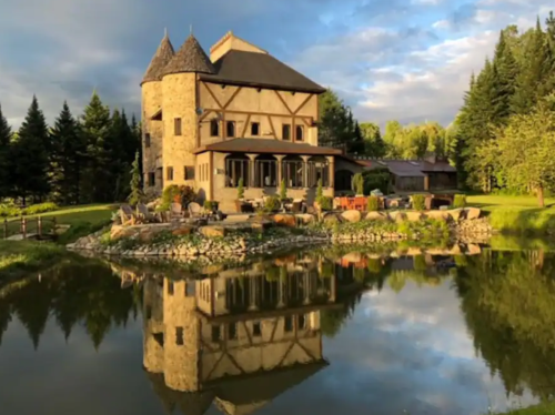 A picturesque stone castle with turrets, surrounded by trees and reflecting in a calm pond under a cloudy sky.