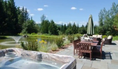 A serene outdoor space featuring a hot tub, dining area, and a pond surrounded by trees and blue skies.