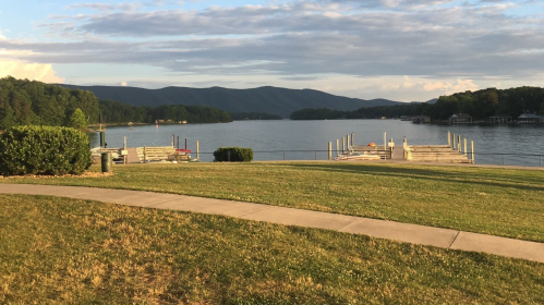 A serene lake view with docks, green grass, and mountains in the background under a partly cloudy sky.