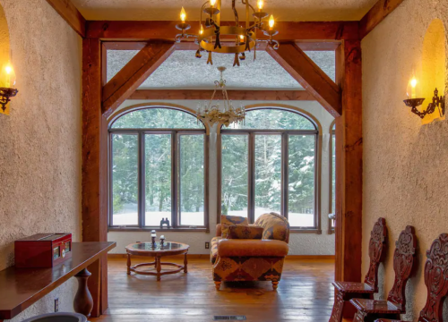 Cozy interior with wooden beams, large windows, and a comfortable chair overlooking a snowy landscape.
