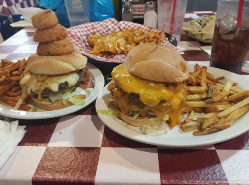 Two large burgers with cheese and toppings, served with fries and a plate of nachos in the background.