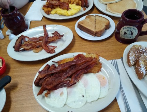 A table set with breakfast plates featuring bacon, eggs, pancakes, toast, and coffee mugs.