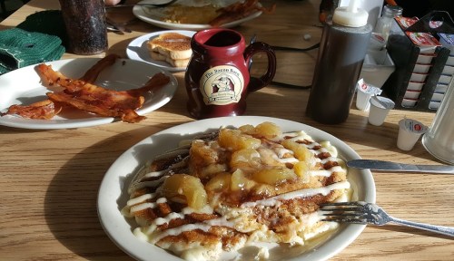 A plate of pancakes topped with apples and syrup, alongside bacon and a coffee mug at a diner table.