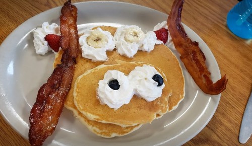 A playful pancake face with whipped cream eyes, banana slices, strawberries, and bacon strips as hair.