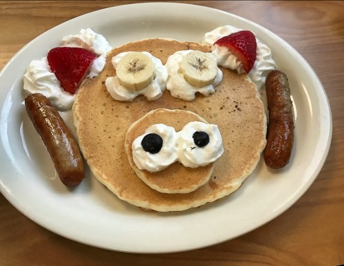 A playful pancake face with banana eyes, whipped cream, strawberries, and sausage on a plate.