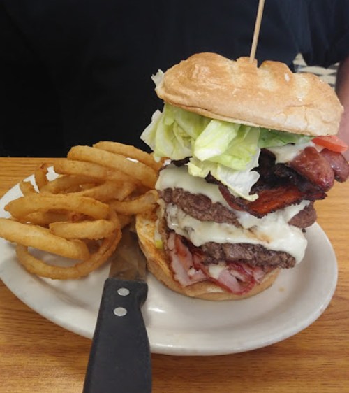 A towering burger with multiple patties, bacon, lettuce, and tomato, served with curly fries on a plate.