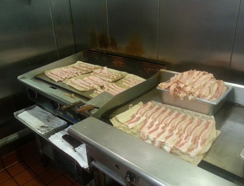 Bacon strips cooking on a griddle in a commercial kitchen, with trays of raw bacon nearby.