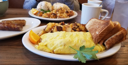 A plate with a fluffy omelette, toast, and a slice of orange, with a side of potatoes and sausage in the background.