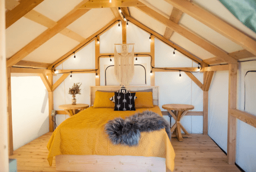 Cozy glamping tent interior with a yellow bedspread, wooden furniture, and string lights.