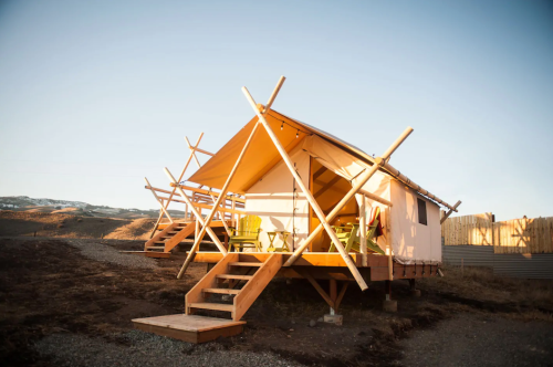 A modern tent cabin on stilts, featuring wooden steps and a deck with green chairs, set against a clear sky.