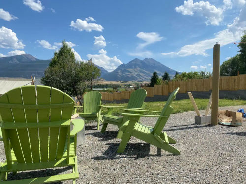 Green Adirondack chairs surround a fire pit, with mountains and a blue sky in the background.
