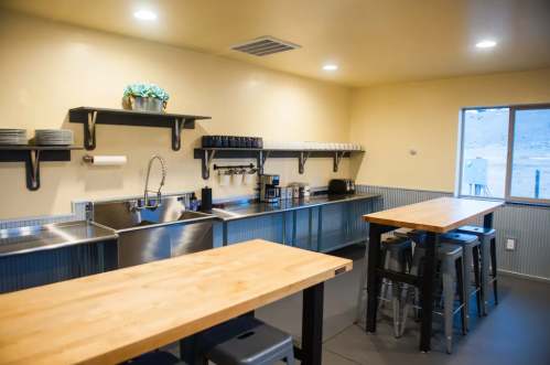 Modern kitchen with stainless steel sink, wooden tables, shelves, and coffee station, featuring a bright, clean design.