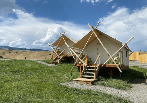 Two glamping tents on wooden platforms, surrounded by green grass and hills under a partly cloudy sky.