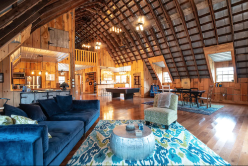 Cozy interior of a barn-style living space with wooden beams, a blue sofa, and a dining area. Bright and inviting atmosphere.